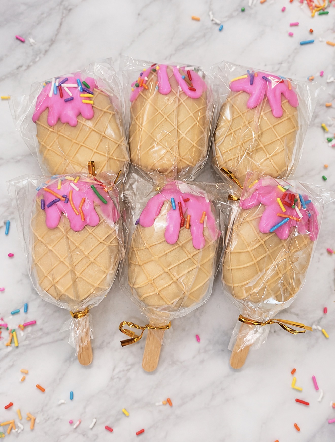 Six vanilla cakesicles with pink drizzle and rainbow sprinkles, individually wrapped with gold ties