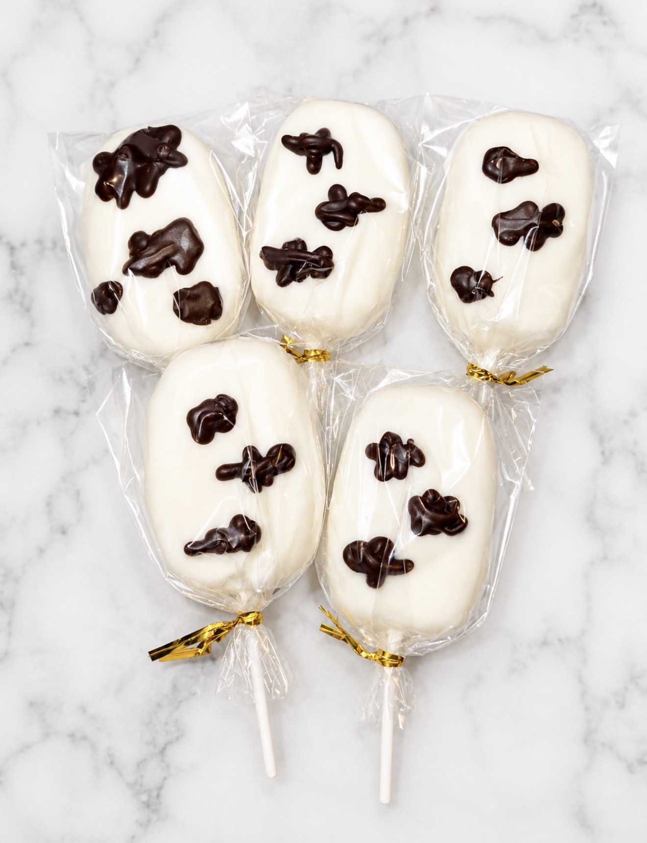 Five white chocolate cakesicles with dark chocolate accents, wrapped with gold ties on marble